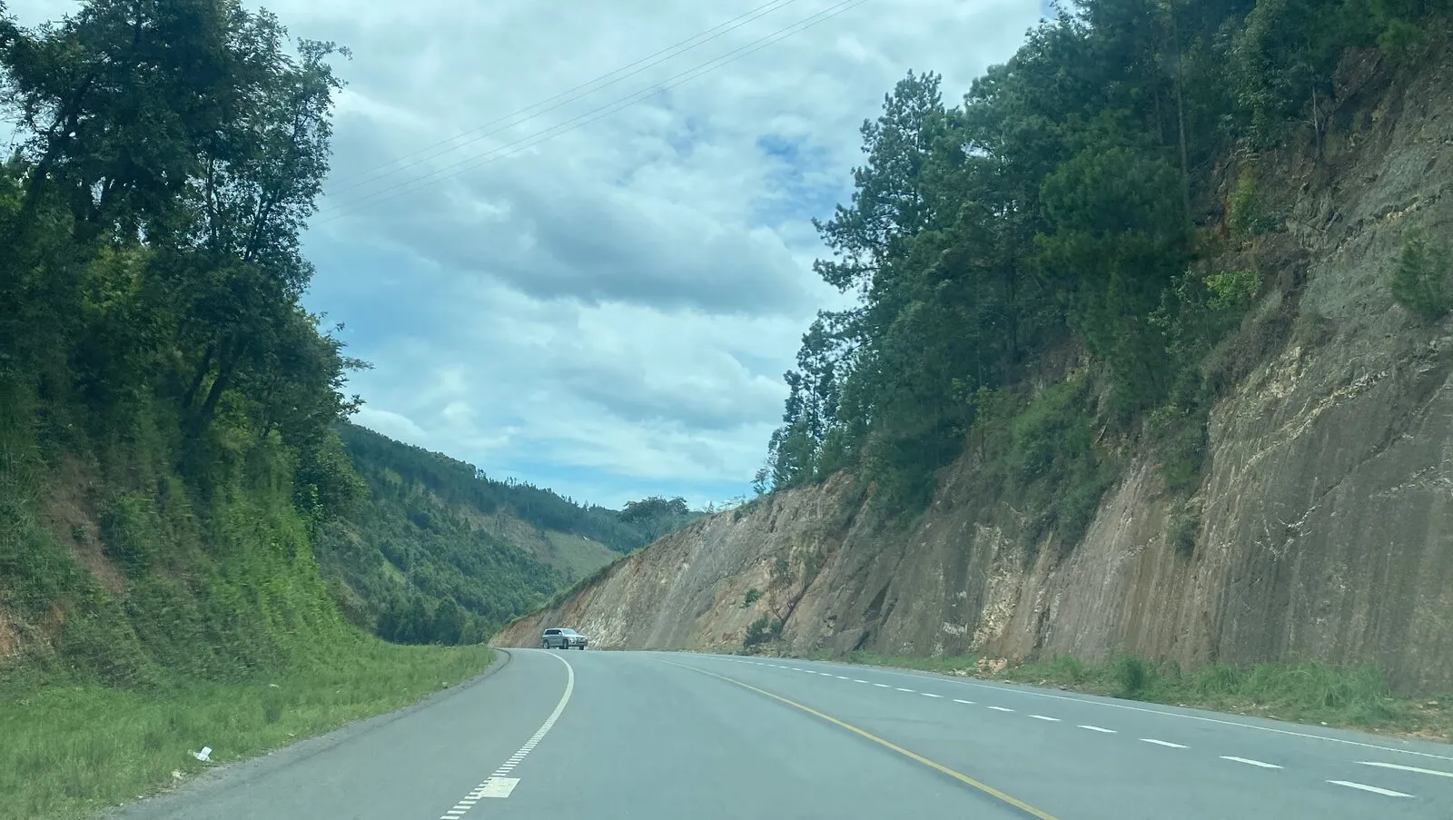 A car driving a winding tarmac road cut through hilly green terrain and rock, on the Bushenyi-to-Kasese road in western Uganda