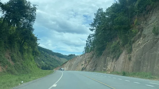 A car driving a winding tarmac road cut through hilly green terrain and rock, on the Bushenyi-to-Kasese road in western Uganda