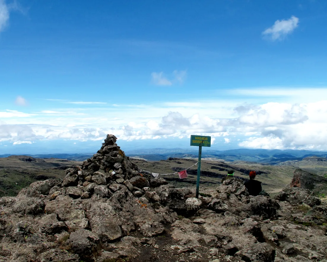 Hikers resting beside the stone summit cairn marking Wagagai Peak, 4,321 metres, on the caldera rim of Mount Elgon
